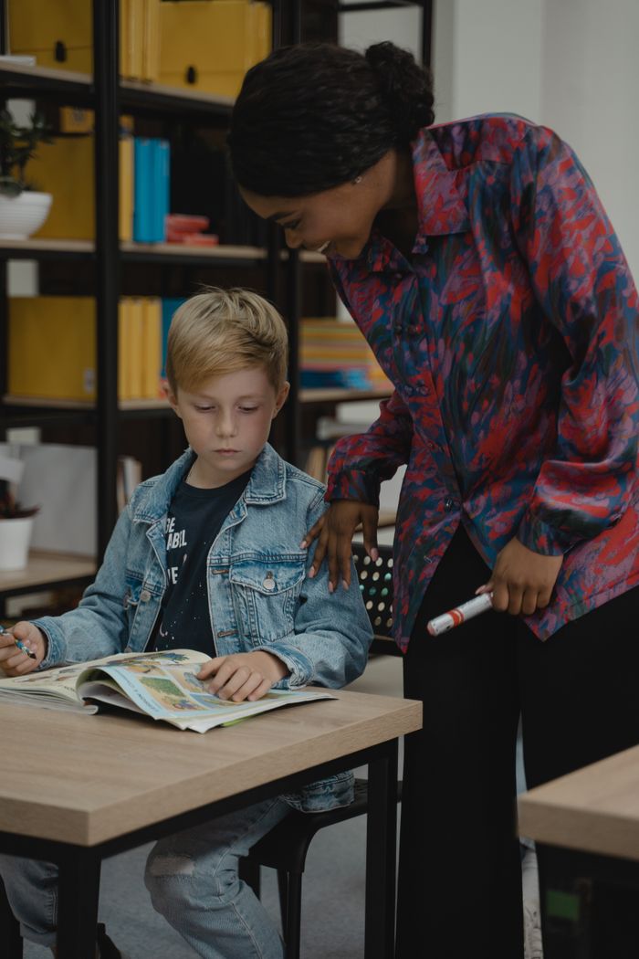 A teacher guides a student reading a book in a classroom setting.