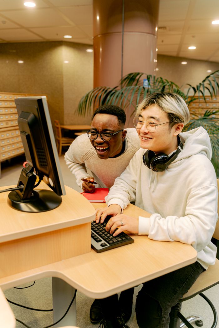 Two students laughing and working together on a computer in a library setting.